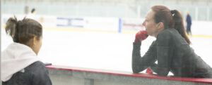 Allison leaning on boards of an ice rink taking a break with her coach in a photo for for the manleywoman skatecast, a figure skating podcast